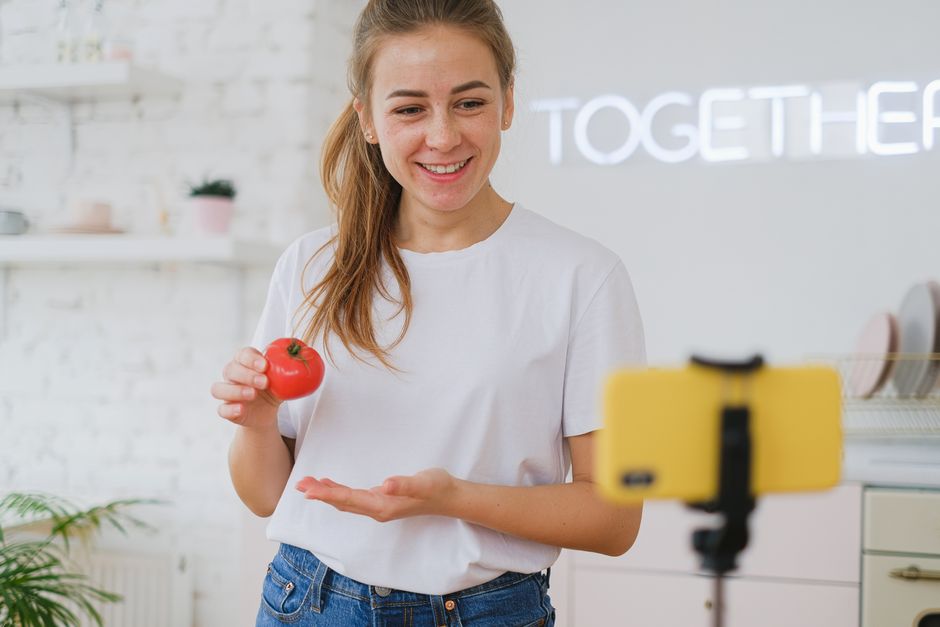 Jovem sorridente segurando um tomate em um ambiente leve e decorado, preparando-se para um vídeo de culinária ou alimentação saudável.