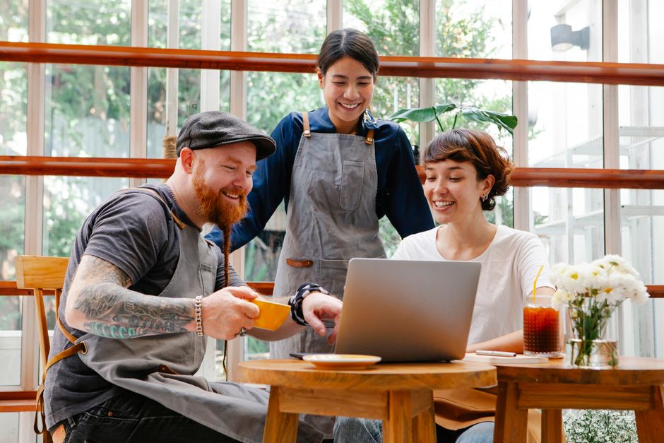Um grupo de amigos trabalhando e conversando de forma descontraída em uma cafeteria com ambiente natural ao fundo, compartilhando ideias e sorrindo enquanto usam um laptop.