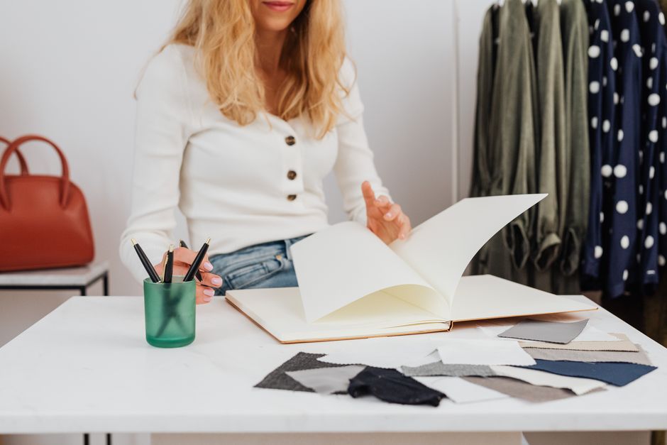 Pessoa analisando amostras de tecidos em um ambiente de moda ou design de moda, com roupas penduradas ao fundo