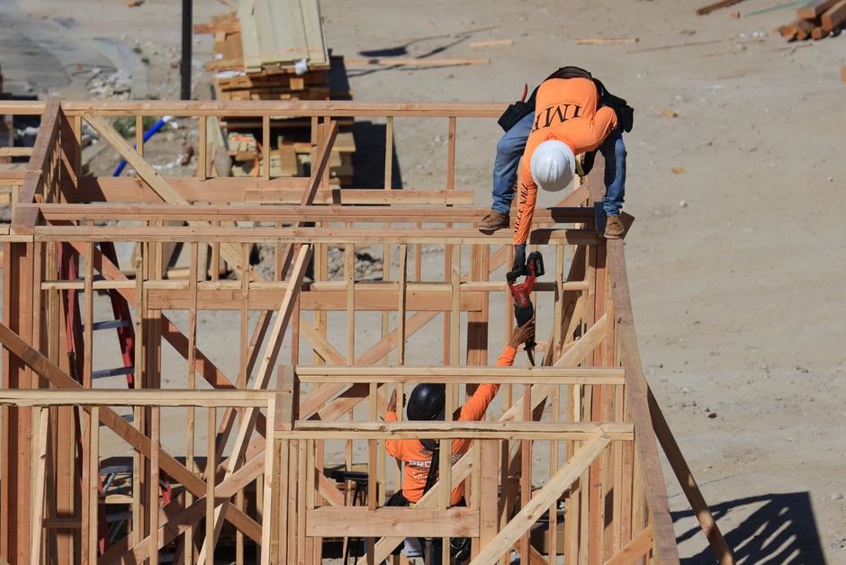 Operários de construção trabalhando na montagem da estrutura de uma edificação de madeira em um canteiro de obras ao ar livre.
