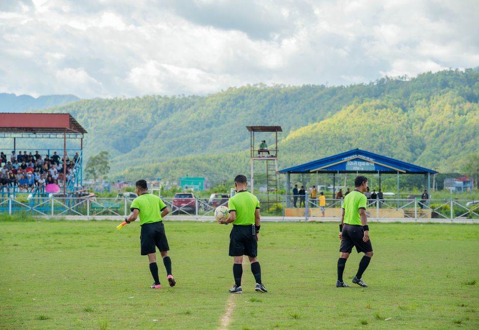 Três árbitros de futebol em campo durante uma partida, com torcedores nas arquibancadas e uma paisagem de colinas verdes ao fundo.