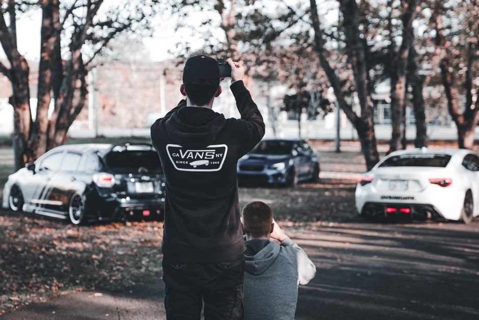 Pessoa registrando carros em um parque, destacando o interesse por automóveis ou atividades ao ar livre relacionadas a veículos.