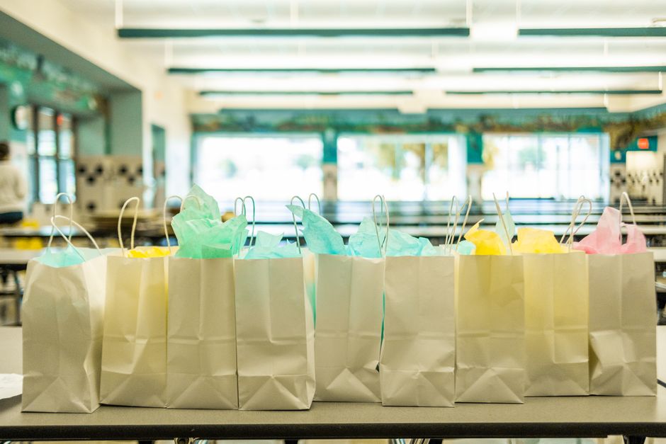 Imagem de sacolas de papel decoradas com papel colorido, em ambiente de salão de festas ou evento infantil, indicando preparação para uma celebração ou entrega de presentes.