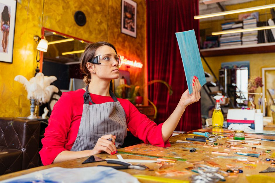 Mulher artesã ou designer analisando uma peça de trabalho em seu ateliê com cores vibrantes e instrumentos de criação ao redor.