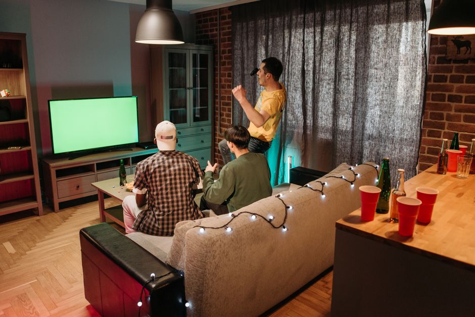 Três amigos se divertindo assistindo a um vídeo em uma sala de estar decorada com luzes e bebidas, celebrando momentos de lazer e amizade.
