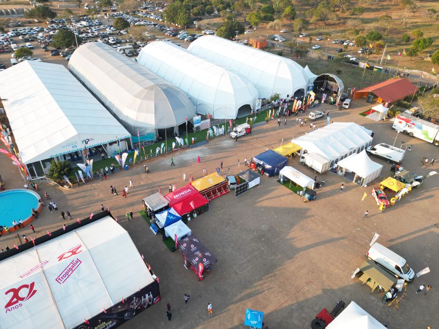 Perspectiva aérea de um evento ao ar livre com grandes tendas coloridas e visitantes, destacando a organização de uma feira ou exposição industrial no campo.