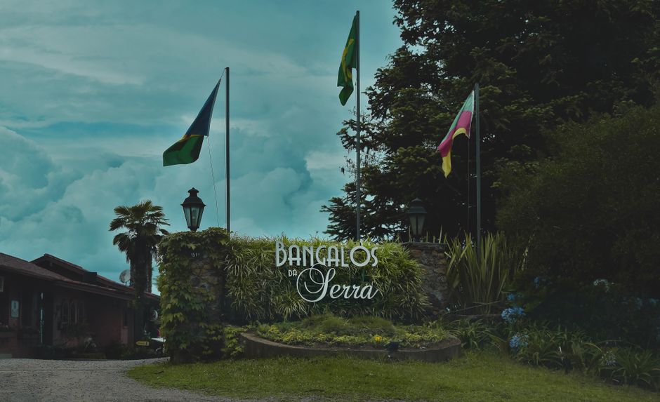 Imagem de entrada de um local chamado 'Bangalos da Serra', com bandeiras e sinalização, situado em um ambiente natural com árvores e plantas ao redor.