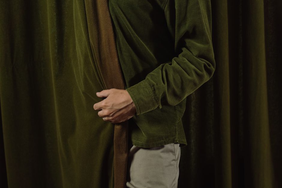 Pessoa se preparando para uma apresentação ou peça teatral, ajustando o roupão em um cenário com cortinas de veludo verde escuro.