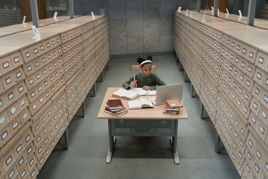 Juventude estudando e pesquisando em uma biblioteca com muitas estantes de fichários, focada no tema de pesquisa acadêmica e organização de estudos.