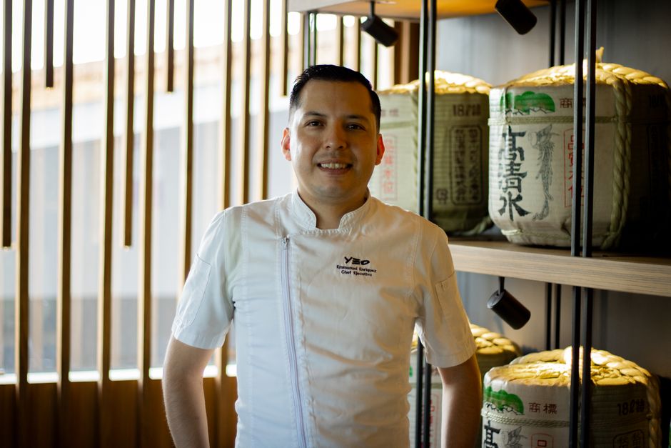 Chef profissional sorridente posando em ambiente de cozinha moderna com ingredientes tradicionais ao fundo.