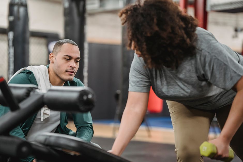 Homem ajudando mulher a fazer exercícios físicos em uma academia, promovendo suporte e orientação fitness.