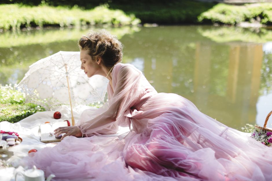 Mulher vestida com um vestido longo rosa claro fazendo um piquenique ao lado de um rio em um ambiente natural e tranquilo.