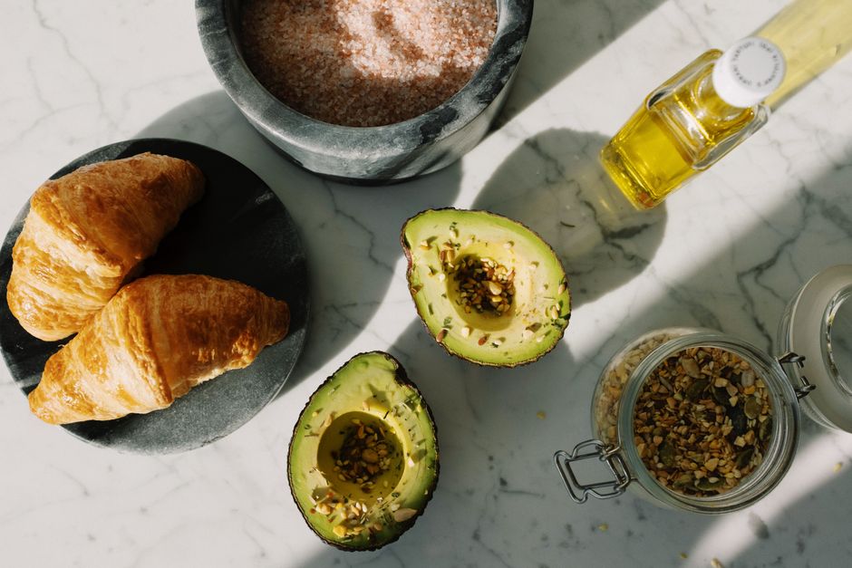 Imagem de café da manhã saudável com pão croissant, abacate fatiado, sementes e óleo de azeite, ideal para artigos sobre alimentação equilibrada ou receitas nutritivas.
