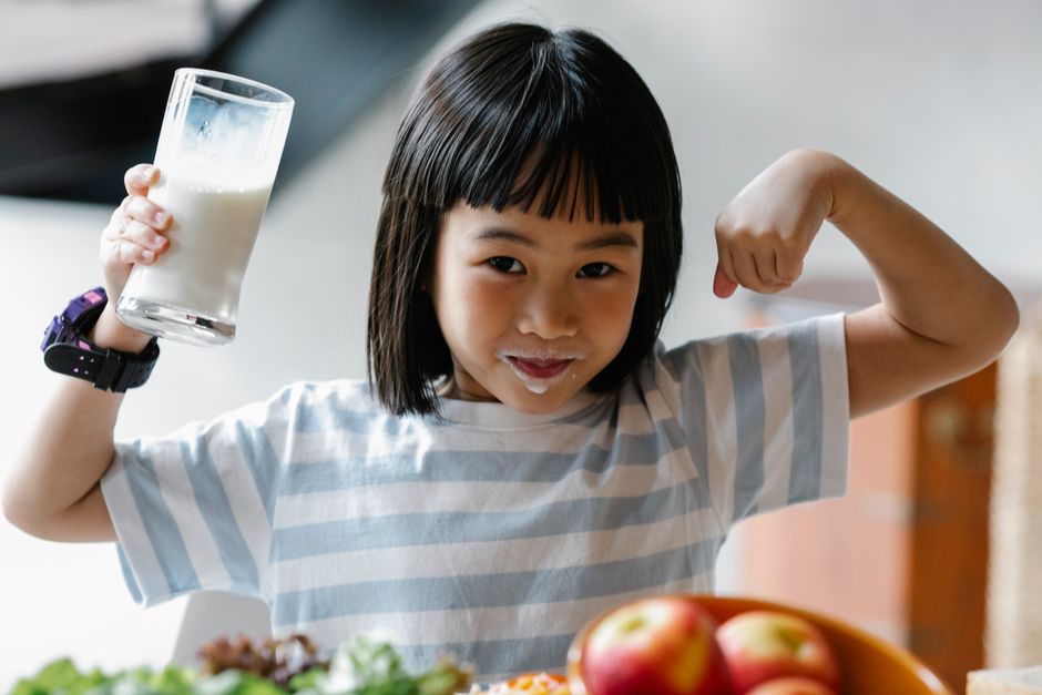 Menina feliz mostrando força após tomar um copo de leite, simbolizando saúde e nutrição infantil.