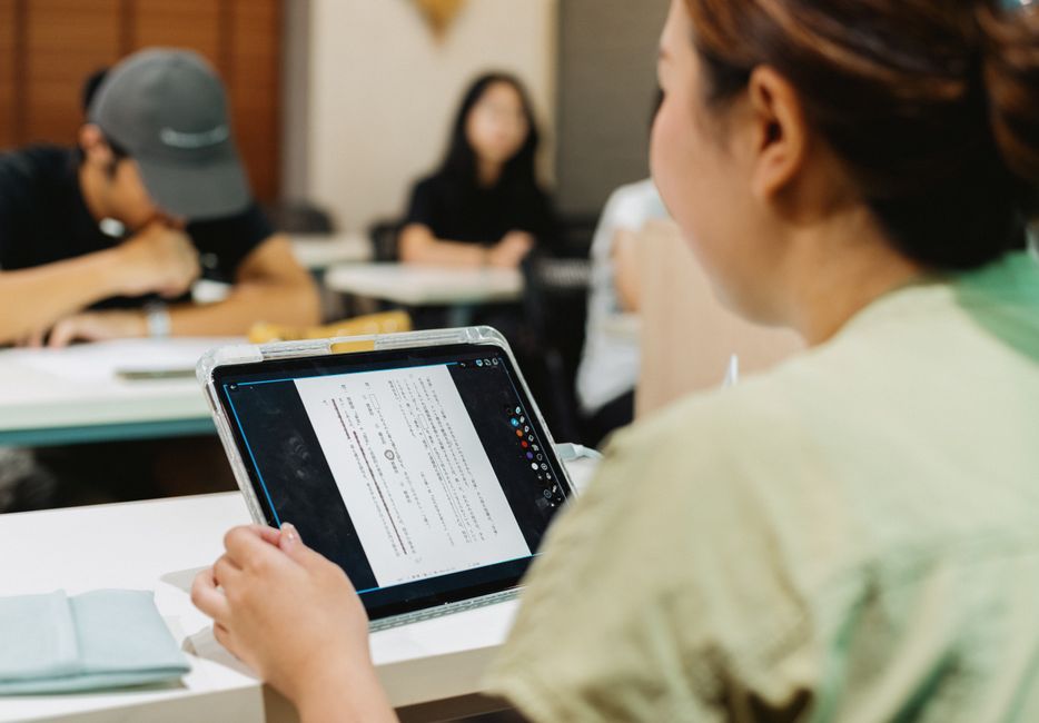 Mulher participando de uma aula ou reunião, utilizando um tablet para leitura ou apresentação, em ambiente corporativo ou educacional.