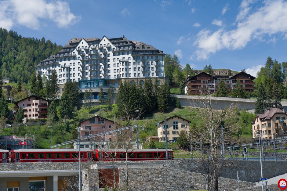 Imagem de uma paisagem montanhosa com edifícios residenciais e um trem vermelho na linha férrea, destacando a integração de natureza e urbanismo em um ambiente de turismo ou moradia de alta qualidade.