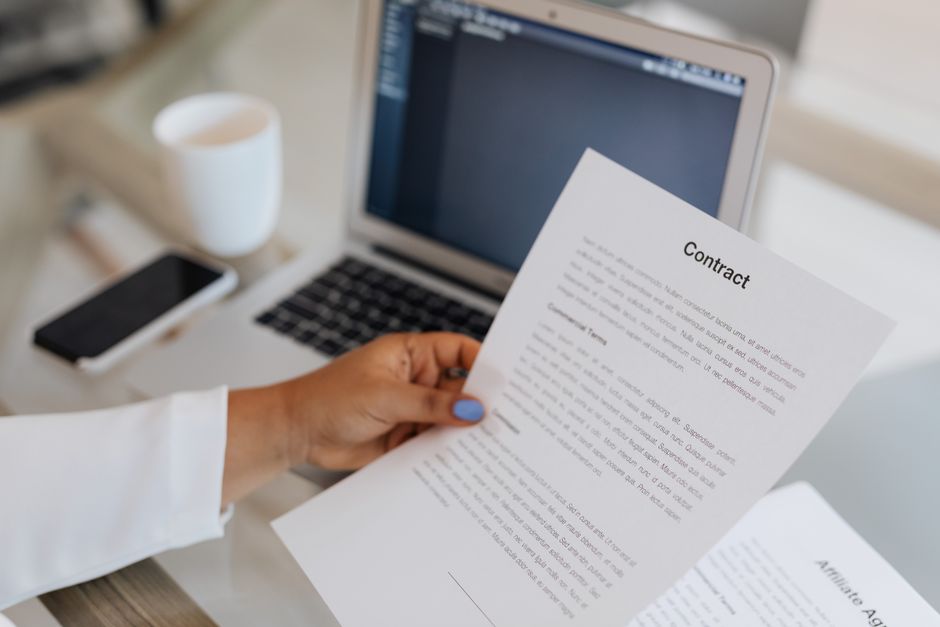 Pessoa revisando um contrato em um ambiente de trabalho com laptop e objetos de escritório ao fundo, para temas de negócios ou direito contratual.