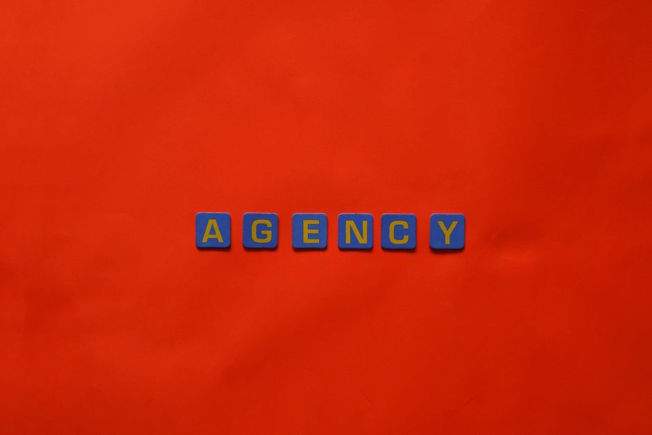 Imagem com blocos de letras formando a palavra 'AGENCY' sobre fundo laranja, representando conceito de agência ou negócios.