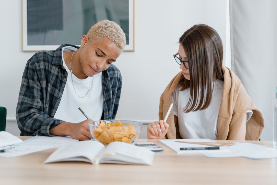 Dois jovens estudantes estudando juntos em uma mesa, com livros, caderno e snacks, em um ambiente de estudo ou sala de aula.