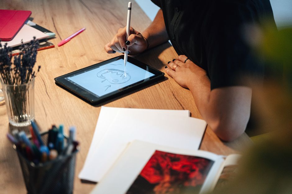 Pessoa usando um tablet para criar um desenho digital em um ambiente de trabalho criativo, com material artístico ao redor.