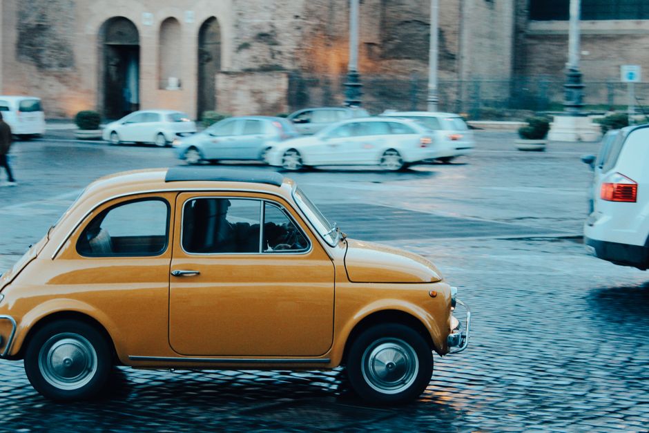 Imagem de um carro clássico amarelo em movimento pelas ruas de uma cidade antiga, destacando-se na cena urbana.
