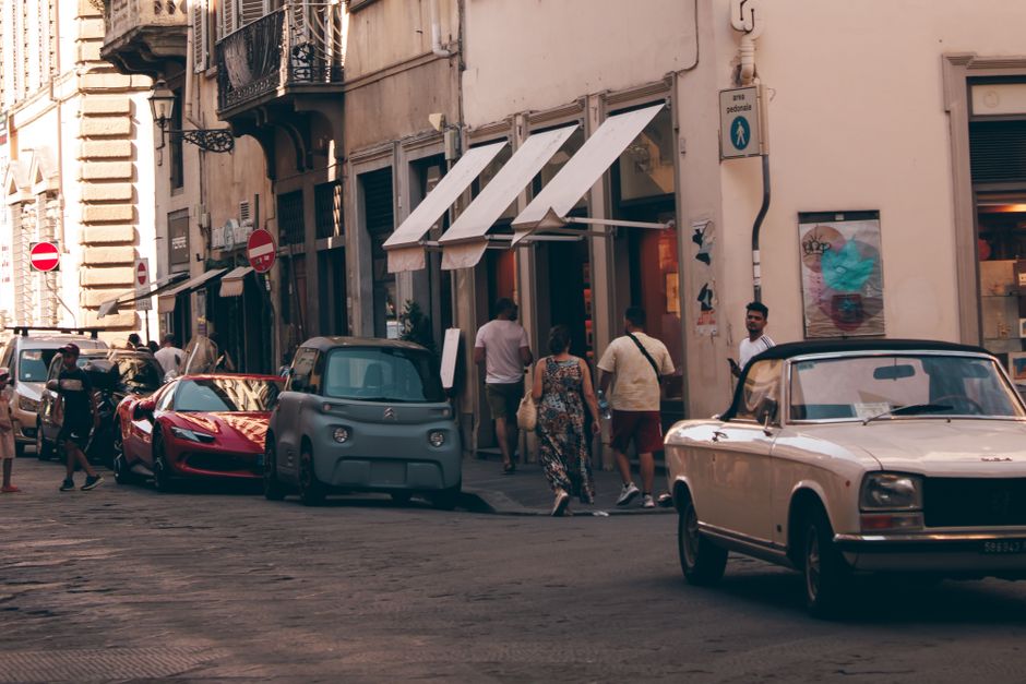 Cena de uma rua urbana movimentada com pedestres entrando em uma loja, veículos clássicos e modernos estacionados ao lado de edifícios antigos, retratando a energia do centro de uma cidade europeia.