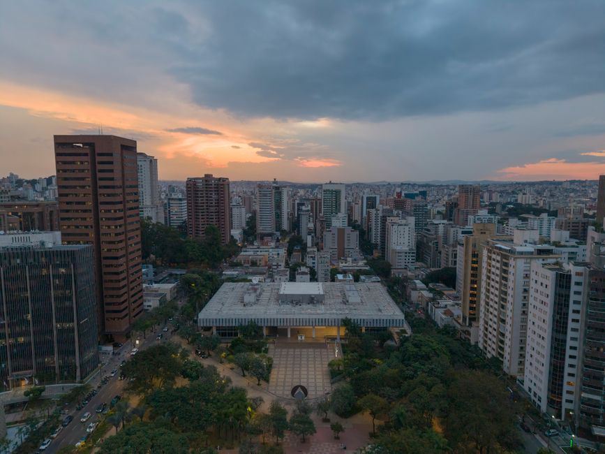 Vista aérea de uma cidade ao entardecer, com prédios altos e céu parcialmente nublado, transmitindo a sensação de urbanismo e movimento.