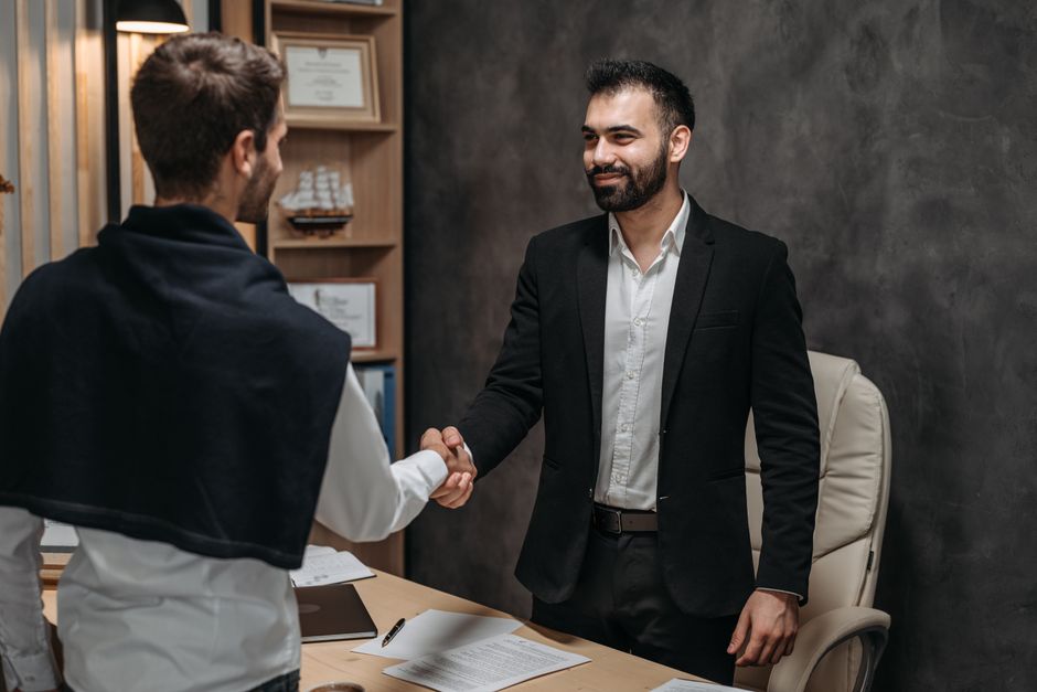 Homem de traje apertando a mão de outro profissional em um ambiente de negócios, simbolizando uma parceria ou acordo corporativo.