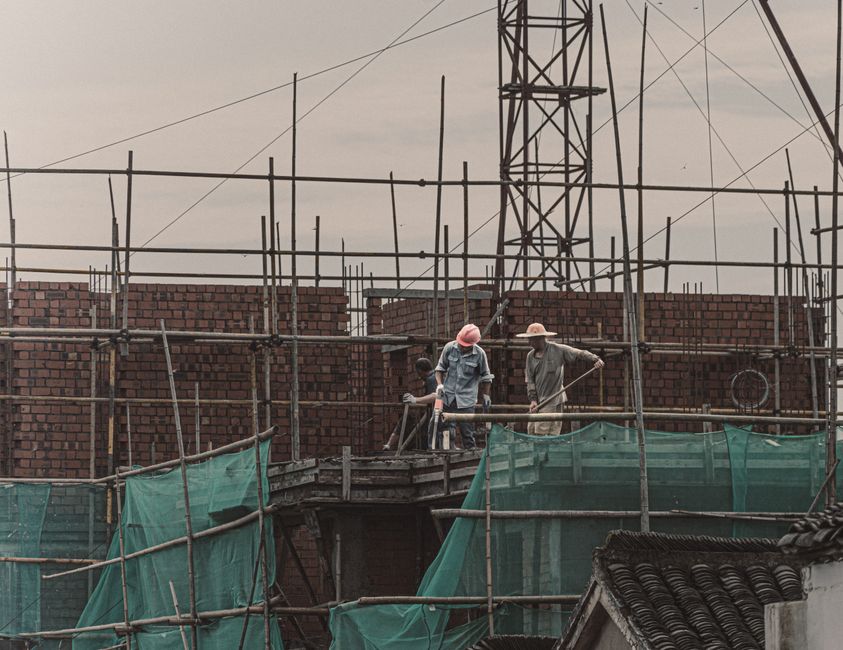 Trabalhadores construindo um prédio com estrutura de madeira e tijolos, durante o dia, em um cenário de obra civil.