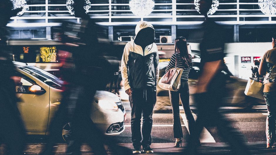 Pessoa esperando no ponto de ônibus em um ambiente urbano movimentado à noite, com movimento de pedestres ao redor.