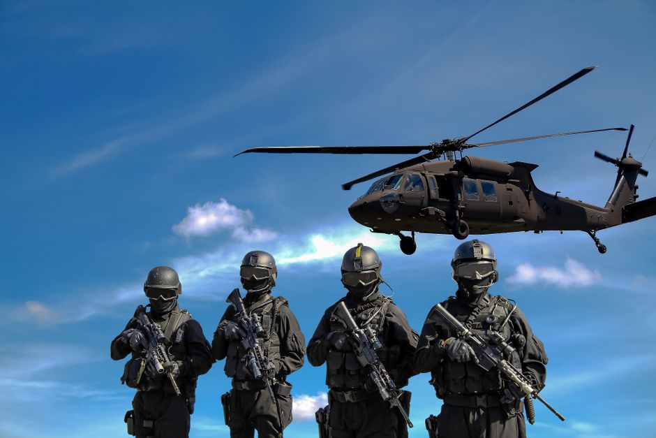 Grupo de soldados equipados com armamentos e traje tático, com um helicóptero em voo ao fundo, em cenário de ação militar.