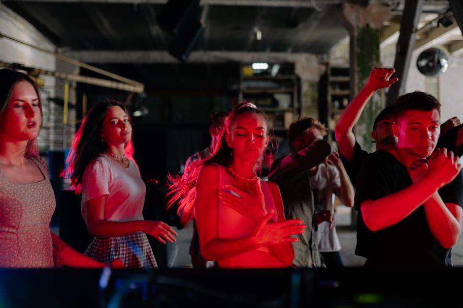 Pessoas dançando e curtindo uma festa ou evento social com iluminação ambiente de cor vermelha, em um espaço com estrutura industrial ao fundo.