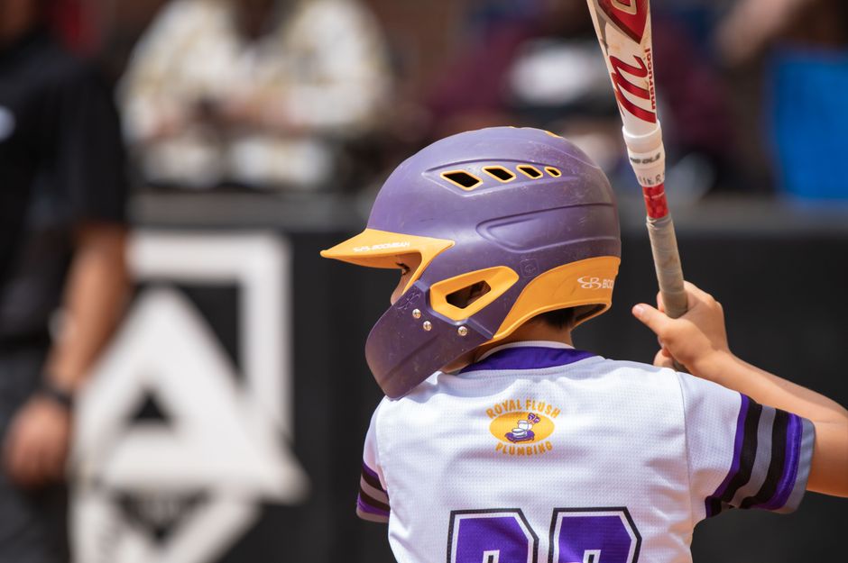 Criança jogando beisebol, preparada para o jogo com capacete e bastão, em um ambiente externo com fundo desfocado.