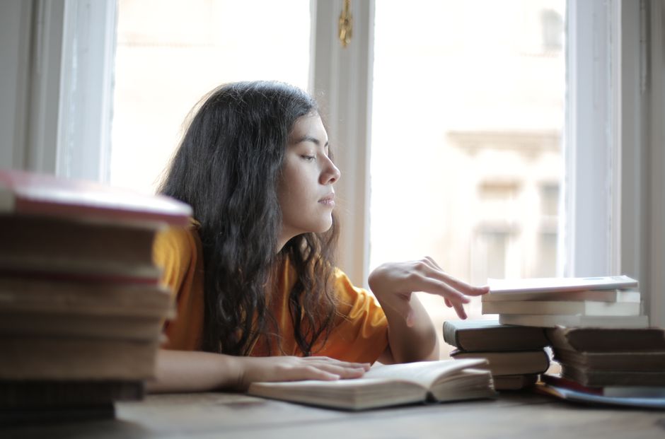 Jovem estudante concentrada estudando com livros e caderno ao lado perto de uma janela que entra luz natural.
