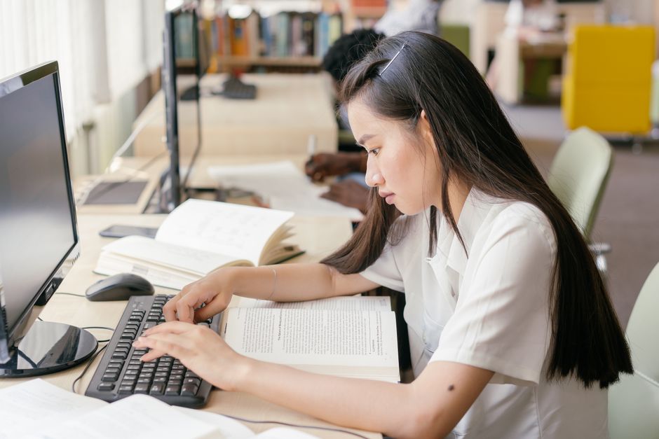 Estudante concentrada estudando em uma biblioteca moderna, rodeada de livros e computadores, com foco em aprendizado e pesquisa.