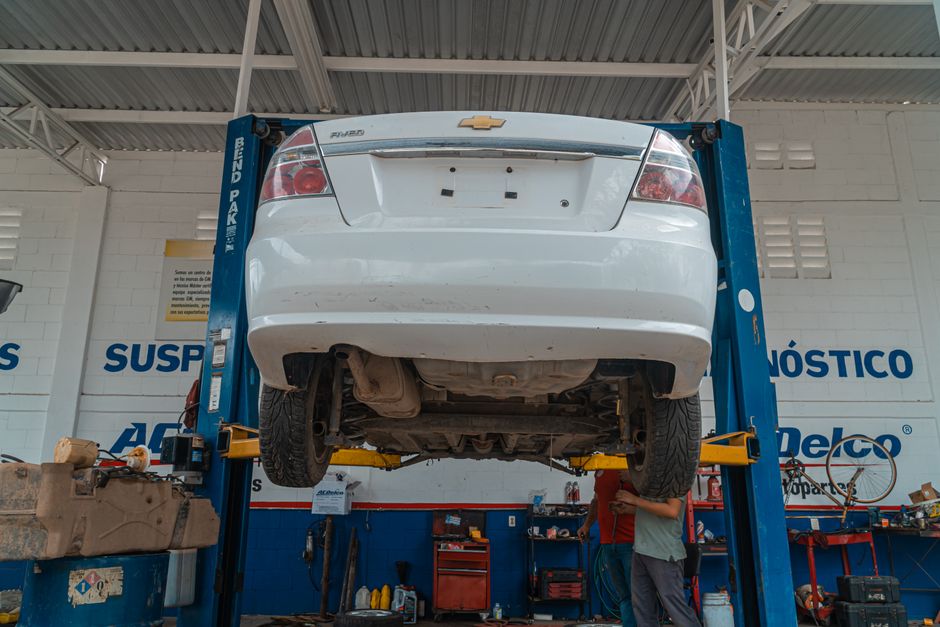 Imagem de um carro branco elevado em uma oficina mecânica, com profissionais realizando manutenção ou reparo na parte inferior do veículo.