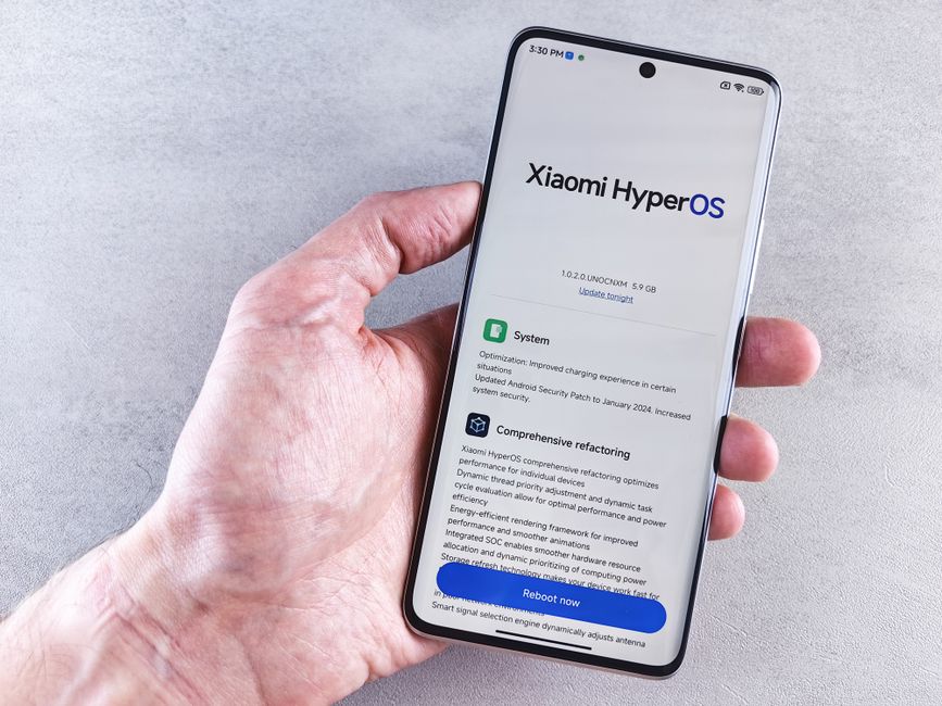 Pessoa segurando um smartphone com a tela exibindo uma atualização do sistema Xiaomi HyperOS, em um ambiente neutro.