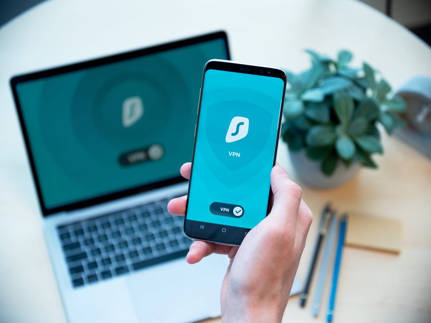Pessoa usando um aplicativo de VPN em um smartphone, com laptop, cadernos e planta no ambiente de trabalho, ilustrando segurança digital e privacidade na internet.