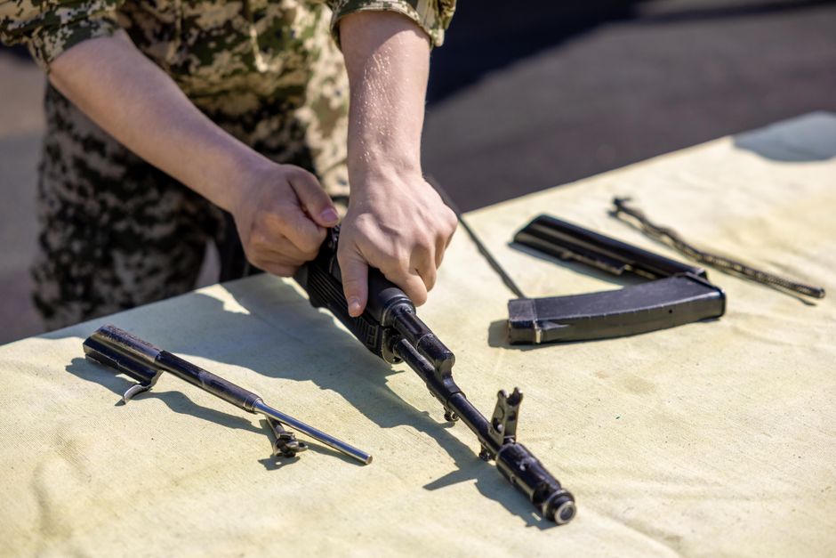 Imagem de uma pessoa manuseando uma arma de fogo com outros itens de armamento sobre uma superfície, sugerindo um contexto militar ou de treinamento de tiro.