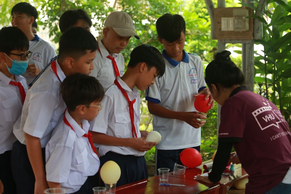 Grupo de estudantes participando de uma atividade escolar ao ar livre, envolvendo preparação de objetos em um ambiente natural.