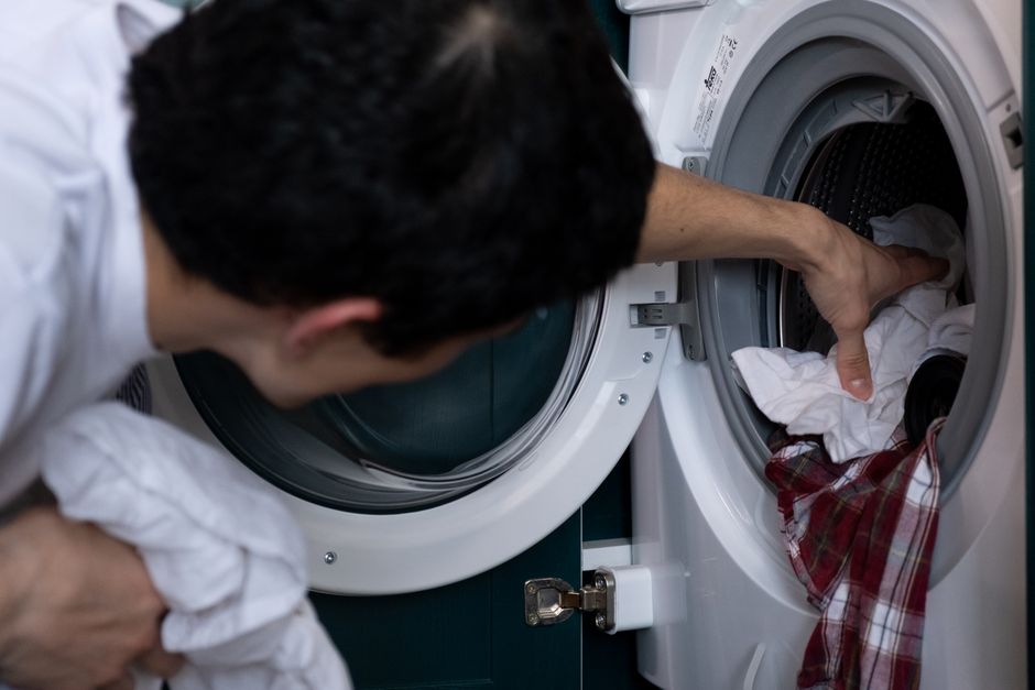 Pessoa carregando roupas em uma máquina de lavar, simbolizando tarefas domésticas ou cuidados com a lavanderia.