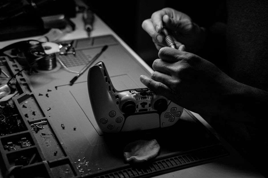 Imagem de uma pessoa consertando um controle de videogame em um ambiente de trabalho técnico ou de hobby, com ferramentas e componentes eletrônicos ao redor.