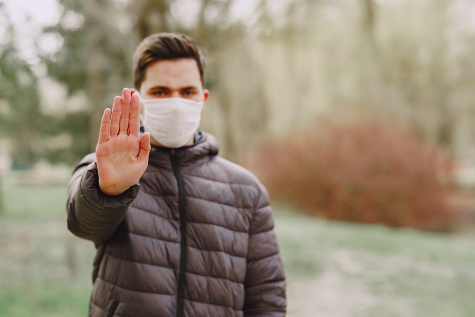 Homem fazendo sinal de parada com máscara facial em ambiente externo, simbolizando cuidados com saúde e prevenção durante a pandemia.