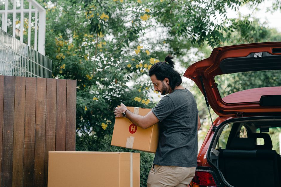 Homem carregando caixas de entrega ao lado de um carro aberto, em um ambiente ao ar livre, simbolizando transporte ou logística.