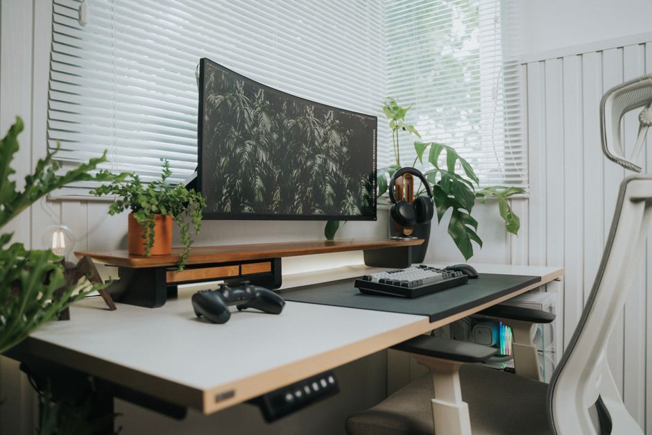 Imagem de um espaço de trabalho em casa equipado com monitor, teclado, videogame, fone de ouvido e plantas, destacando um ambiente de produtividade e lazer.
