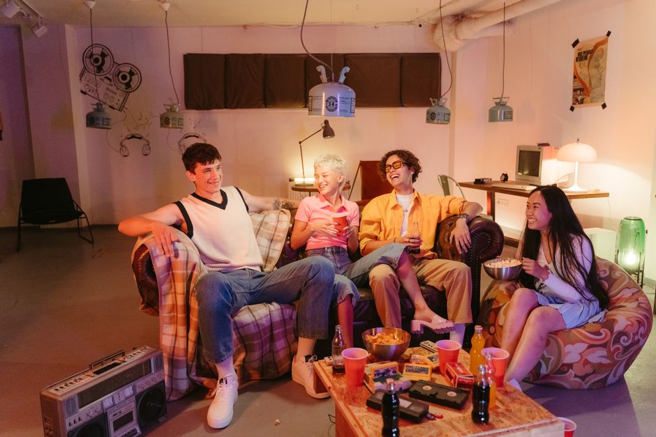 Grupo de amigos jovens sorrindo e conversando em uma festa descontraída em um ambiente interno com iluminação quente.