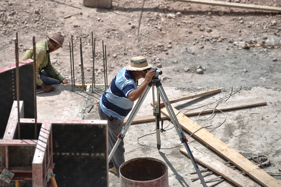 Homem usando um nível de precisão em um canteiro de obras ao lado de um cinegrafista com um tripé, ambos trabalhando em um projeto de construção.