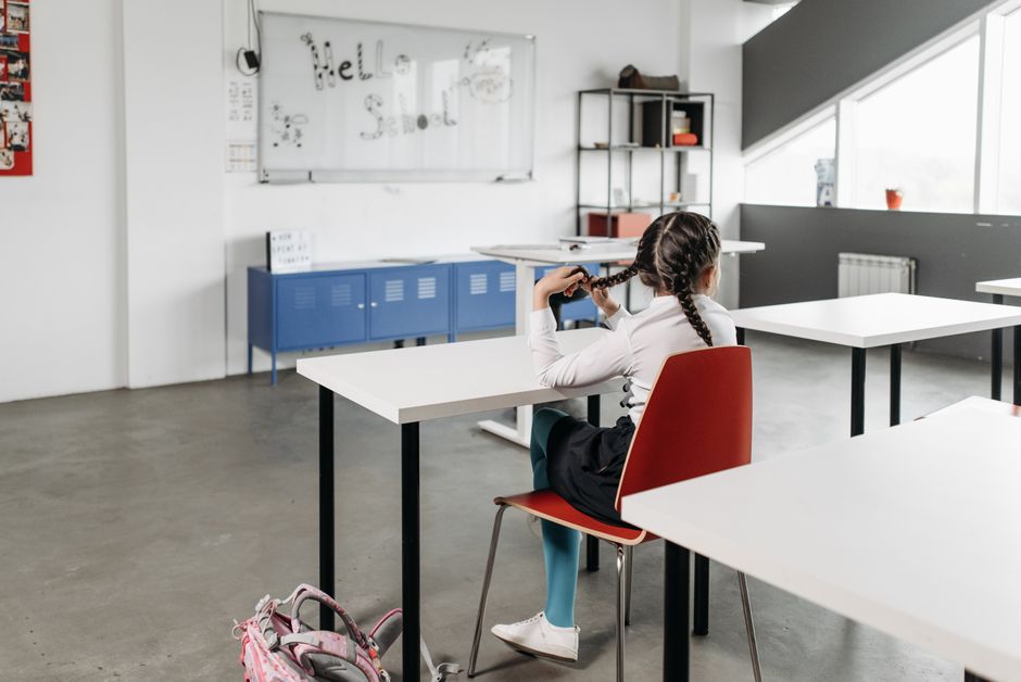 Criança assistindo aula em sala de aula moderna, com mesa e quadro branco ao fundo.