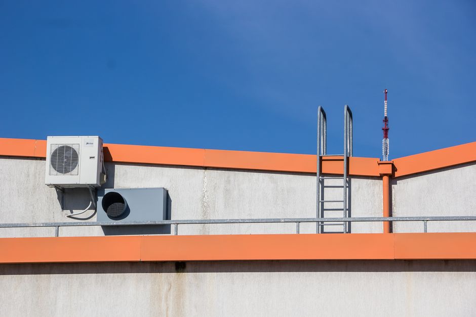 Imagem de uma cobertura de edifício com elementos técnicos como escada, ventilação e sistema de ar-condicionado contra um céu claro, representando manutenção predial ou infraestrutura urbana.
