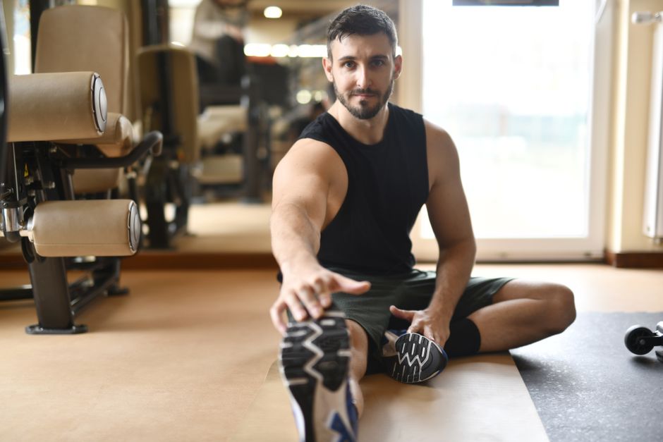 Homem realizando alongamento ou exercício de flexibilidade em ambiente de academia ou espaço de treino, evidenciando saúde e bem-estar.